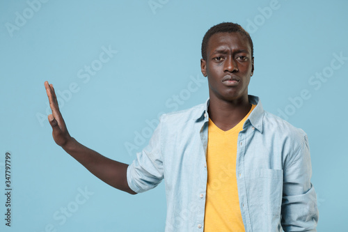 Dissatisfied young african american man guy in casual shirt, yellow t-shirt posing isolated on blue wall background. People lifestyle concept. Mock up copy space. Showing stop gesture with palm aside.