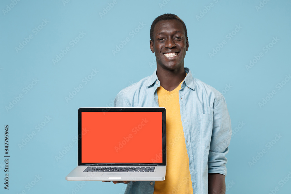 Smiling young african american man guy in casual shirt yellow t-shirt posing isolated on blue background. People lifestyle concept. Mock up copy space. Hold laptop pc computer with blank empty screen. Smiling young african american man guy in casual shirt yellow t-shirt posing isolated on blue background. People lifestyle concept. Mock up copy space. Hold laptop pc computer with blank empty screen.