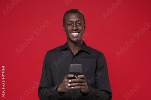 Smiling young african american man guy in classic black shirt posing isolated on red background studio portrait. People lifestyle concept. Mock up copy space. Using mobile phone, typing sms message. © ViDi Studio