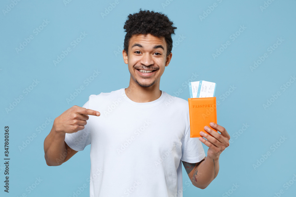 Smiling young african american guy in casual white t-shirt traveling abroad isolated on blue wall background. Air flight journey concept. Mock up copy space. Pointing index finger on passport tickets. Smiling young african american guy in casual white t-shirt traveling abroad isolated on blue wall background. Air flight journey concept. Mock up copy space. Pointing index finger on passport tickets.