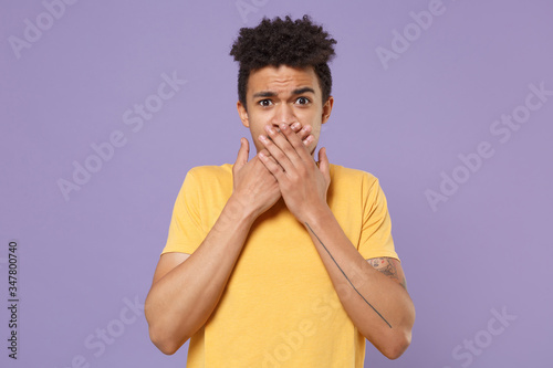 Concerned young african american guy in casual yellow t-shirt posing isolated on pastel violet wall background studio portrait. People lifestyle concept. Mock up copy space. Covering mouth with hands.