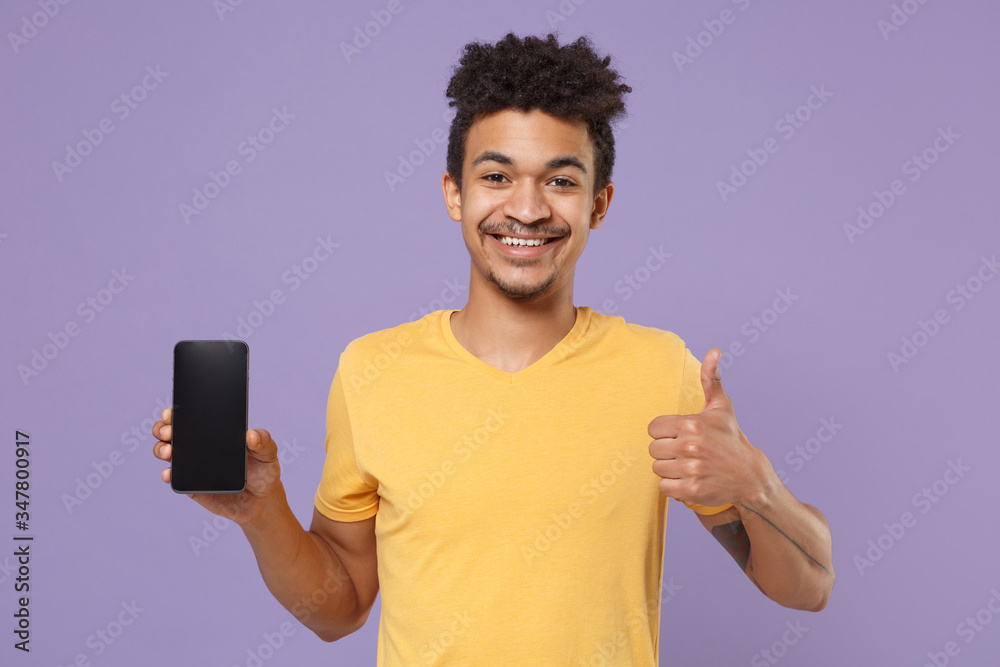 Smiling young african american guy in yellow t-shirt posing isolated on violet background. People lifestyle concept. Mock up copy space. Hold mobile phone with blank empty screen, showing thumb up. Smiling young african american guy in yellow t-shirt posing isolated on violet background. People lifestyle concept. Mock up copy space. Hold mobile phone with blank empty screen, showing thumb up.
