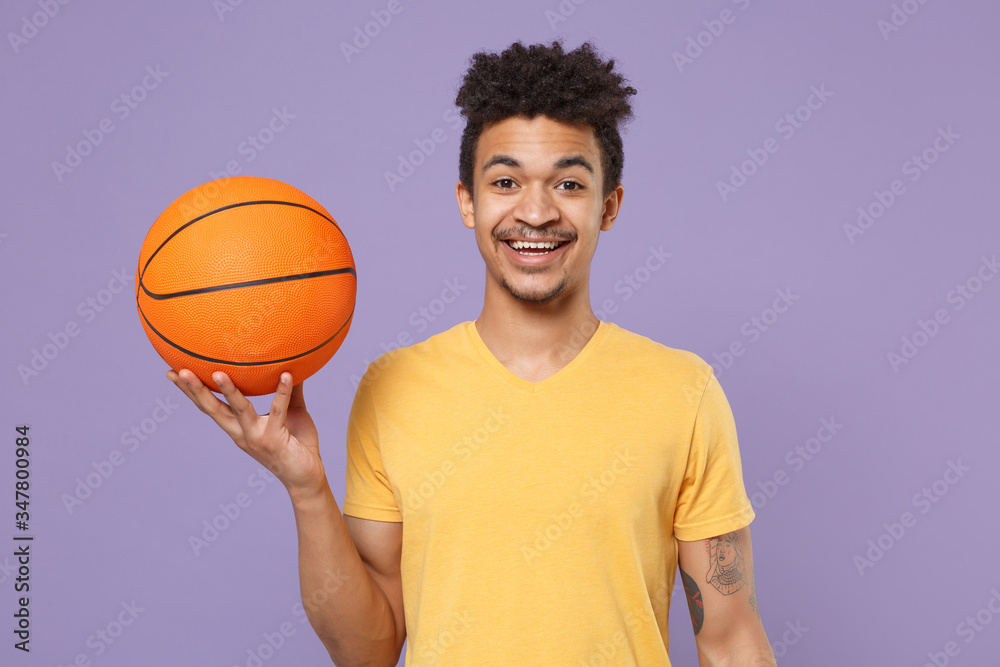 Smiling young african american guy basketball player in casual yellow t-shirt posing isolated on pastel violet background. People emotions, sport leisure lifestyle concept. Play basketball hold ball.