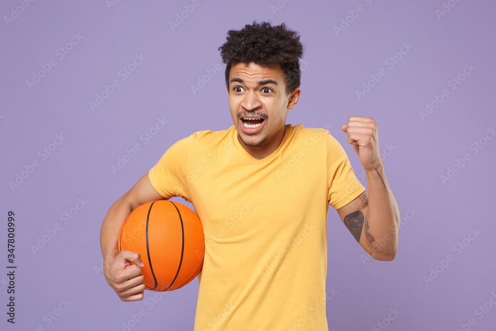 Worried young african american guy basketball player in yellow t-shirt isolated on pastel violet background. People emotions, sport leisure lifestyle concept. Play basketball hold ball clenching fist.