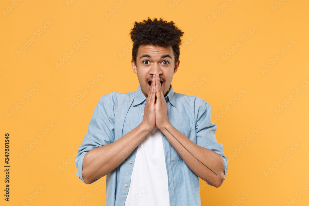 Shocked young african american guy in casual blue shirt posing isolated on yellow wall background studio portrait. People emotions lifestyle concept. Mock up copy space. Covering mouth with hands. Shocked young african american guy in casual blue shirt posing isolated on yellow wall background studio portrait. People emotions lifestyle concept. Mock up copy space. Covering mouth with hands.