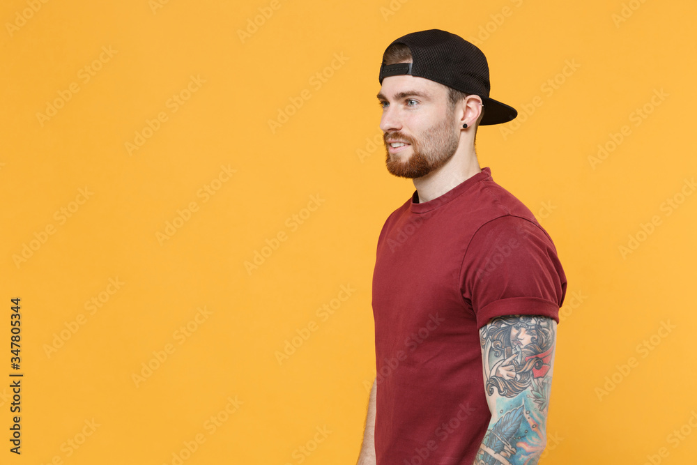 Side view of attractive young bearded tattooed man guy in casual t-shirt, black cap posing isolated on yellow background studio portrait. People lifestyle concept. Mock up copy space. Looking aside. Side view of attractive young bearded tattooed man guy in casual t-shirt, black cap posing isolated on yellow background studio portrait. People lifestyle concept. Mock up copy space. Looking aside.