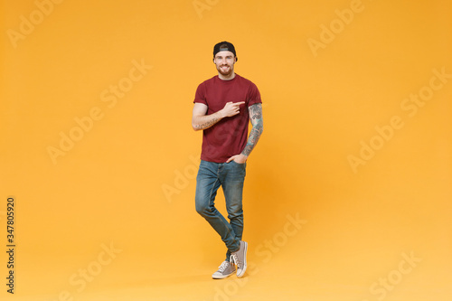Smiling young bearded tattooed man guy in casual t-shirt black cap posing isolated on yellow wall background studio portrait. People lifestyle concept. Mock up copy space. Pointing index finger aside.