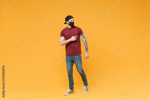 Young tattooed man guy in casual t-shirt cap black face mask posing isolated on yellow background studio portrait. People emotions lifestyle concept. Mock up copy space. Pointing index finger aside.
