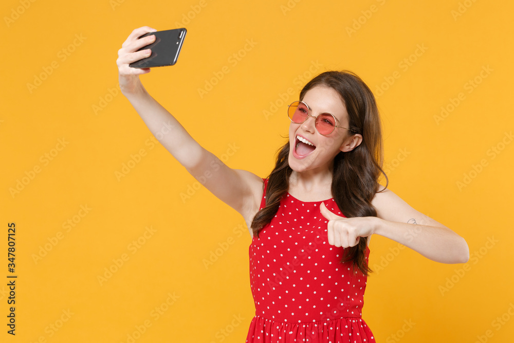 Cheerful brunette woman girl in red summer dress, eyeglasses posing isolated on yellow background. People lifestyle concept. Mock up copy space. Doing selfie shot on mobile phone showing thumb up. Cheerful brunette woman girl in red summer dress, eyeglasses posing isolated on yellow background. People lifestyle concept. Mock up copy space. Doing selfie shot on mobile phone showing thumb up.