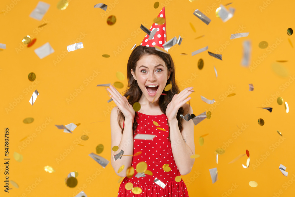 Excited young woman girl in red summer dress birthday hat isolated on yellow wall background studio portrait. Birthday holiday concept. Mock up copy space. Celebrating with confetti, spreading hands.