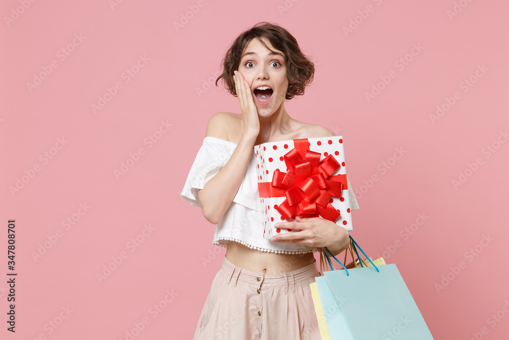 Excited woman in summer clothes isolated on pink background. Shopping discount sale concept. Mock up copy space. Hold package bag with purchases red present box with gift ribbon bow put hand on cheek. Excited woman in summer clothes isolated on pink background. Shopping discount sale concept. Mock up copy space. Hold package bag with purchases red present box with gift ribbon bow put hand on cheek.