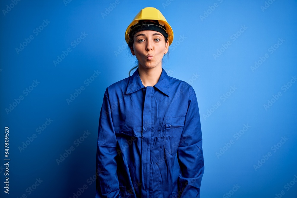 Young beautiful worker woman with blue eyes wearing security helmet and uniform making fish face with lips, crazy and comical gesture. Funny expression.