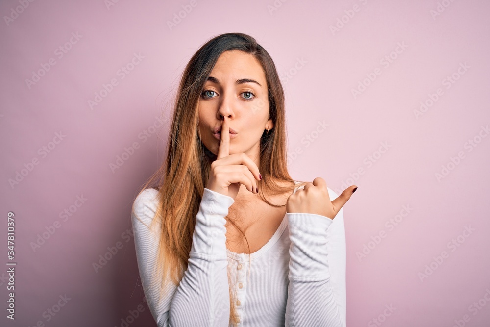Young beautiful blonde woman with blue eyes wearing white t-shirt over pink background asking to be quiet with finger on lips pointing with hand to the side. Silence and secret concept. Young beautiful blonde woman with blue eyes wearing white t-shirt over pink background asking to be quiet with finger on lips pointing with hand to the side. Silence and secret concept.