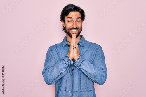 Young handsome hispanic bohemian man wearing hippie style over pink background praying with hands together asking for forgiveness smiling confident.