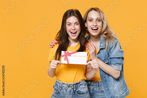 Two excited young women girls friends in casual t-shirts denim clothes posing isolated on yellow wall background studio portrait. People lifestyle concept. Mock up copy space. Hold gift certificate.