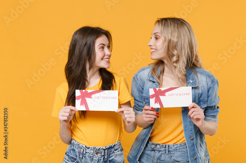Two smiling young women girls friends in casual t-shirts denim clothes posing isolated on yellow background. People lifestyle concept. Mock up copy space. Hold gift certificates looking at each other.