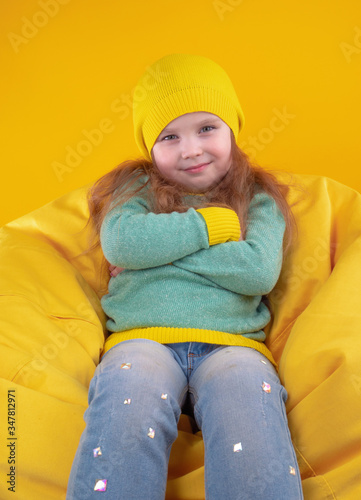 Happy girl in trendy clothes looking up while chilling on soft bean bag against yellow background