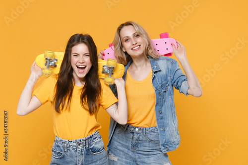 Two cheerful young women girls friends in casual t-shirts denim clothes isolated on yellow background studio portrait. People sincere emotions lifestyle concept. Mock up copy space. Hold skateboards.