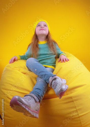 Full body happy girl in trendy clothes looking up while chilling on soft bean bag against yellow background