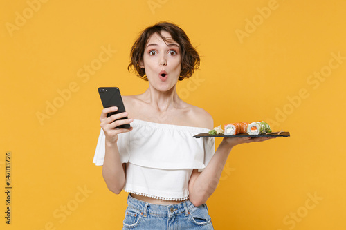 Young woman girl in casual clothes hold in hand makizushi sushi roll served on black plate japanese food using mobile cell phone isolated on yellow background studio portrait. People lifestyle concept