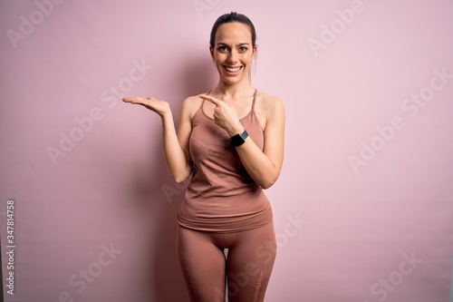 Young blonde fitness woman wearing sport workout clothes over pink isolated background amazed and smiling to the camera while presenting with hand and pointing with finger.