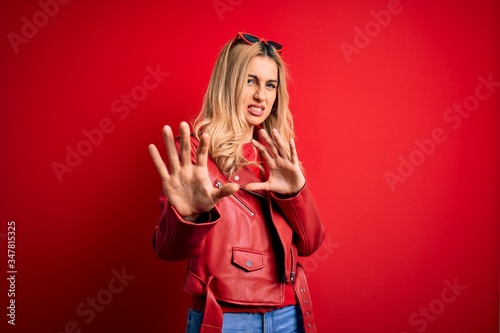 Young beautiful blonde woman wearing casual jacket standing over isolated red background disgusted expression, displeased and fearful doing disgust face because aversion reaction. With hands raised