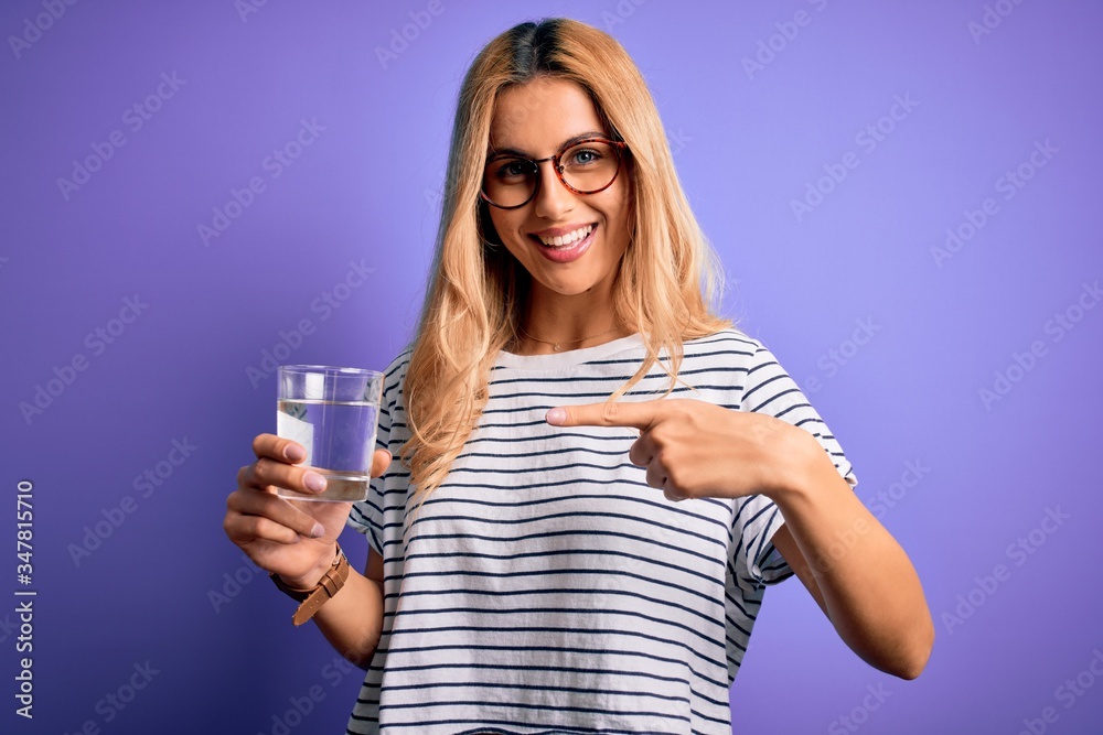 Young blonde healthy woman wearing glasses drinking glass of water over purple background very happy pointing with hand and finger Young blonde healthy woman wearing glasses drinking glass of water over purple background very happy pointing with hand and finger