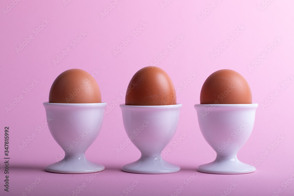 three brown eggs in white eggs cups before pink background three brown eggs in white eggs cups before pink background