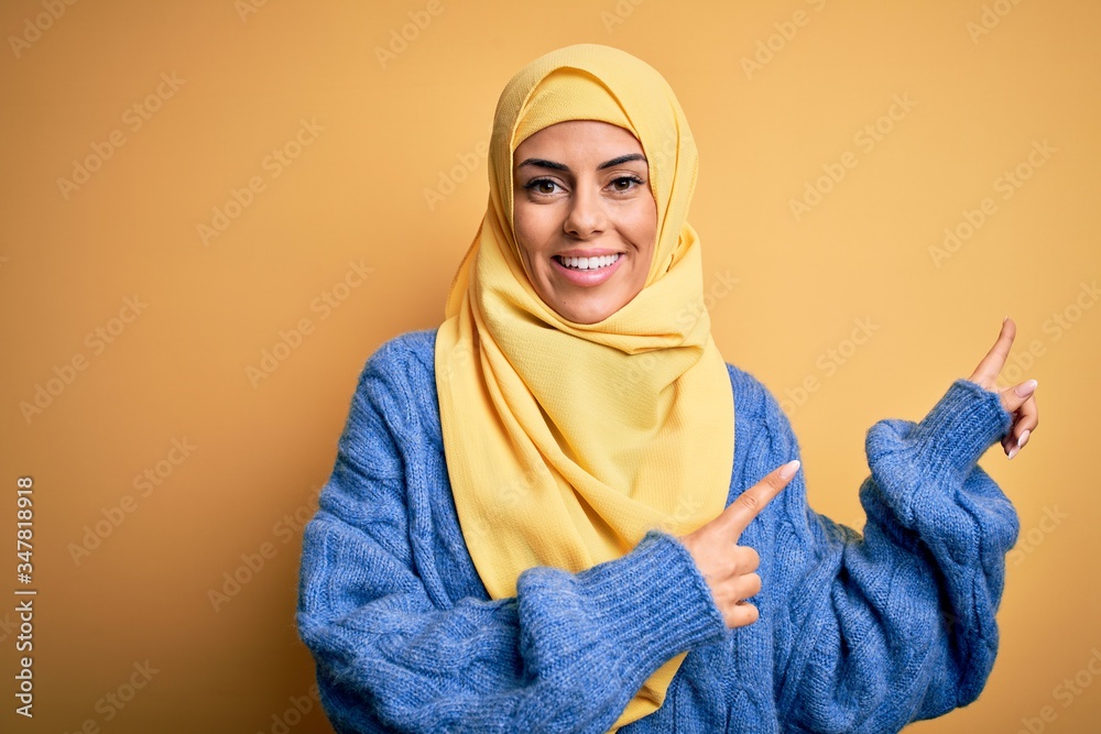 Young beautiful brunette muslim woman wearing arab hijab over isolated yellow background smiling and looking at the camera pointing with two hands and fingers to the side. Young beautiful brunette muslim woman wearing arab hijab over isolated yellow background smiling and looking at the camera pointing with two hands and fingers to the side.
