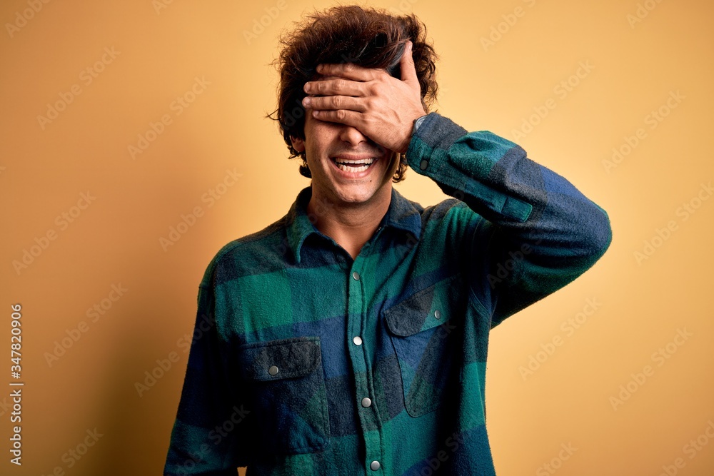 Young handsome man wearing casual shirt standing over isolated yellow background smiling and laughing with hand on face covering eyes for surprise. Blind concept.