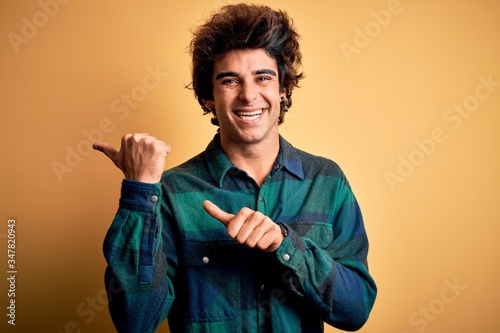 Young handsome man wearing casual shirt standing over isolated yellow background Pointing to the back behind with hand and thumbs up, smiling confident photo