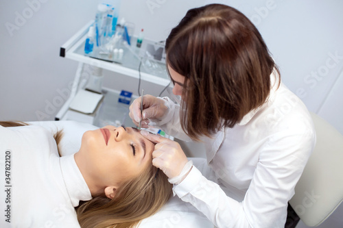 A young woman lies and master in a white uniform does eyebrow makeup in a beauty salon. The use of permanent makeup on brows. The wizard works with eyebrows. Semi-permanent makeup.