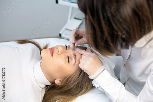 A young woman lies and master in a white uniform does eyebrow makeup in a beauty salon. The use of permanent makeup on brows. Wizard works with eyebrows. Semi-permanent makeup. Girl master at work.