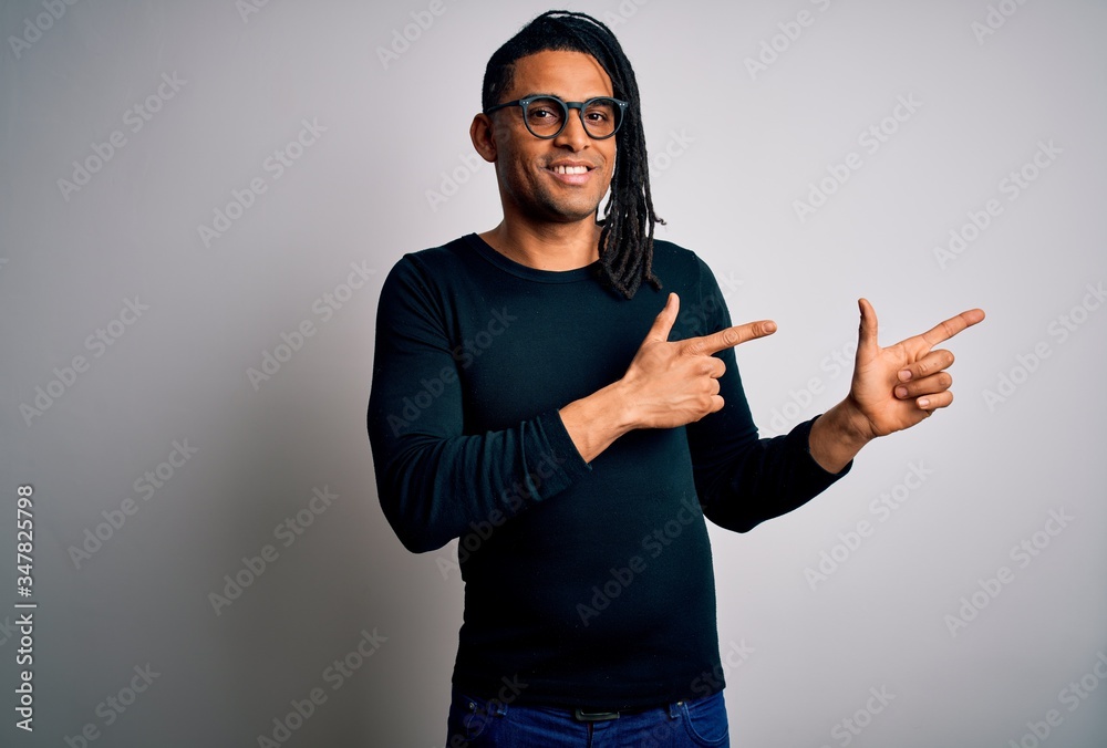 Young handsome african american man with dreadlocks wearing casual t-shirt and glasses smiling and looking at the camera pointing with two hands and fingers to the side. Young handsome african american man with dreadlocks wearing casual t-shirt and glasses smiling and looking at the camera pointing with two hands and fingers to the side.