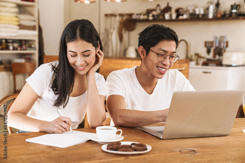 Image of multicultural couple making notes in planner and using laptop
