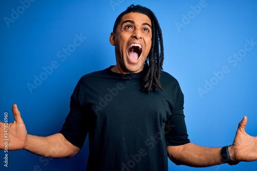 Young handsome african american afro man with dreadlocks wearing casual t-shirt crazy and mad shouting and yelling with aggressive expression and arms raised. Frustration concept.