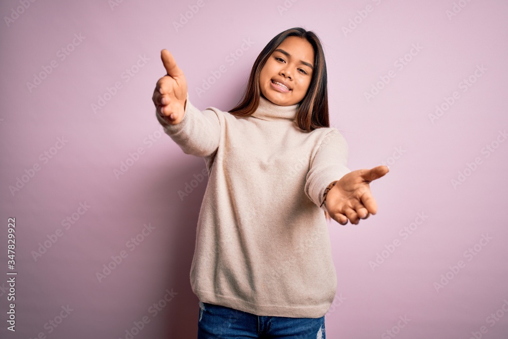 Young beautiful asian girl wearing casual turtleneck sweater over isolated pink background looking at the camera smiling with open arms for hug. Cheerful expression embracing happiness.