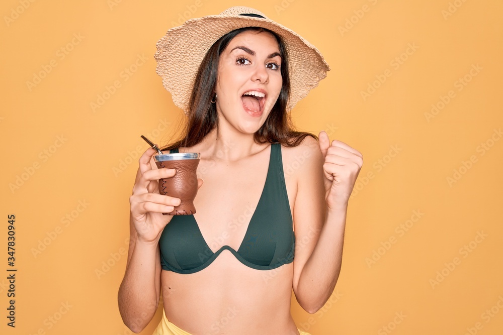 Young beautiful girl wearing swimwear bikini and summer hat drinking traditional mate drink screaming proud and celebrating victory and success very excited, cheering emotion Young beautiful girl wearing swimwear bikini and summer hat drinking traditional mate drink screaming proud and celebrating victory and success very excited, cheering emotion