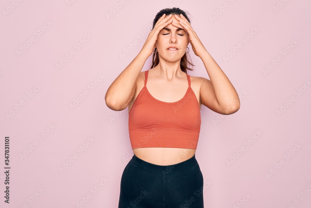 Young beautiful fitness woman wearing sport excersie clothes over pink background suffering from headache desperate and stressed because pain and migraine. Hands on head.