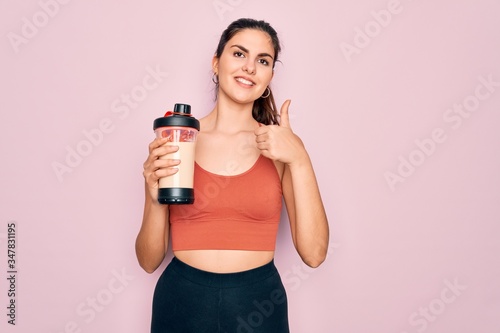 Young beautiful fitness woman wearing sport excersie clothes drinking protein shake happy with big smile doing ok sign, thumb up with fingers, excellent sign © Krakenimages.com