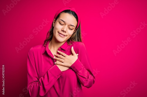 Young beautiful brunette girl wearing casual shirt standing over isolated pink background smiling with hands on chest with closed eyes and grateful gesture on face. Health concept.