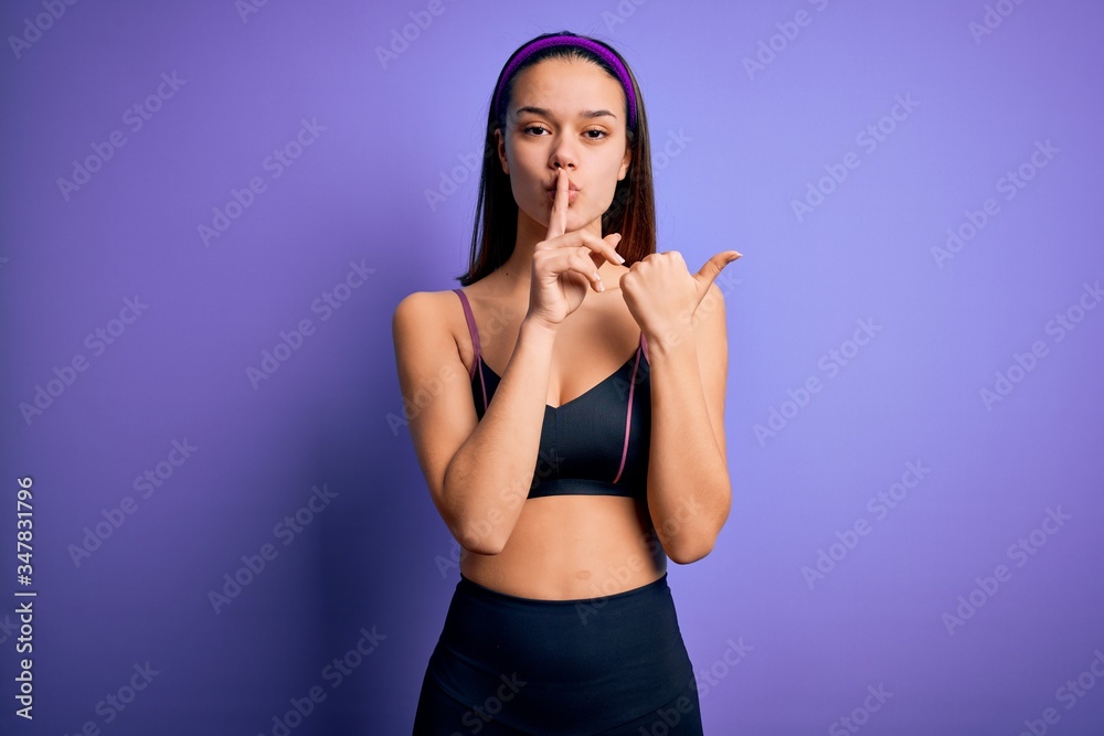 Fototapeta premium Young beautiful sporty girl doing sport wearing sportswear over isolated purple background asking to be quiet with finger on lips pointing with hand to the side. Silence and secret concept.