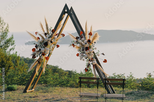 Original wedding arch on top of the mountain with white and red flowers at sunset. Romantic ceremony in the style of boho