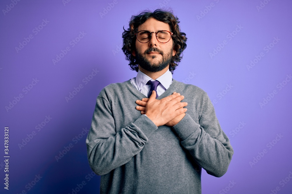 Handsome businessman with beard wearing tie and glasses standing over purple background smiling with hands on chest with closed eyes and grateful gesture on face. Health concept. Handsome businessman with beard wearing tie and glasses standing over purple background smiling with hands on chest with closed eyes and grateful gesture on face. Health concept.