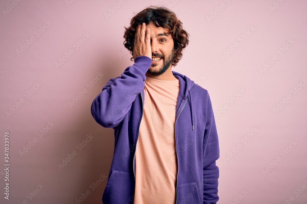 Young handsome sporty man with beard wearing casual sweatshirt over pink background covering one eye with hand, confident smile on face and surprise emotion. Young handsome sporty man with beard wearing casual sweatshirt over pink background covering one eye with hand, confident smile on face and surprise emotion.