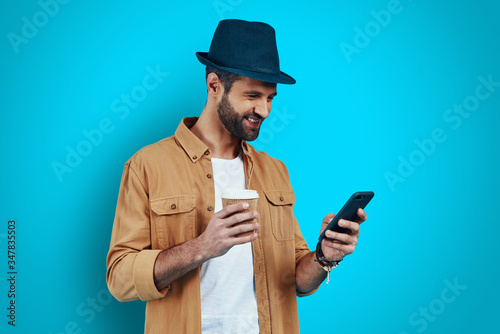 Handsome young man smiling and using smart phone while standing against blue background © G-Stock Faces
