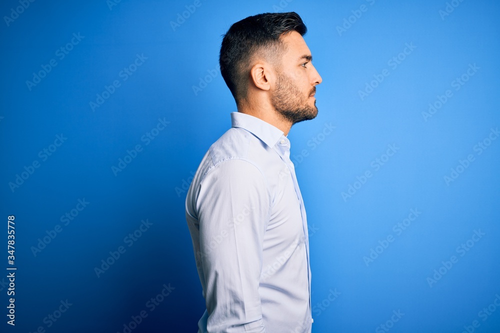 Young handsome man wearing elegant shirt standing over isolated blue background looking to side, relax profile pose with natural face with confident smile.