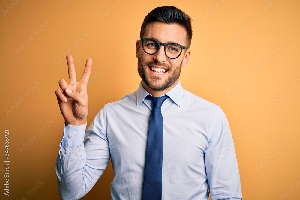 Young handsome businessman wearing tie and glasses standing over yellow background showing and pointing up with fingers number two while smiling confident and happy. Young handsome businessman wearing tie and glasses standing over yellow background showing and pointing up with fingers number two while smiling confident and happy.