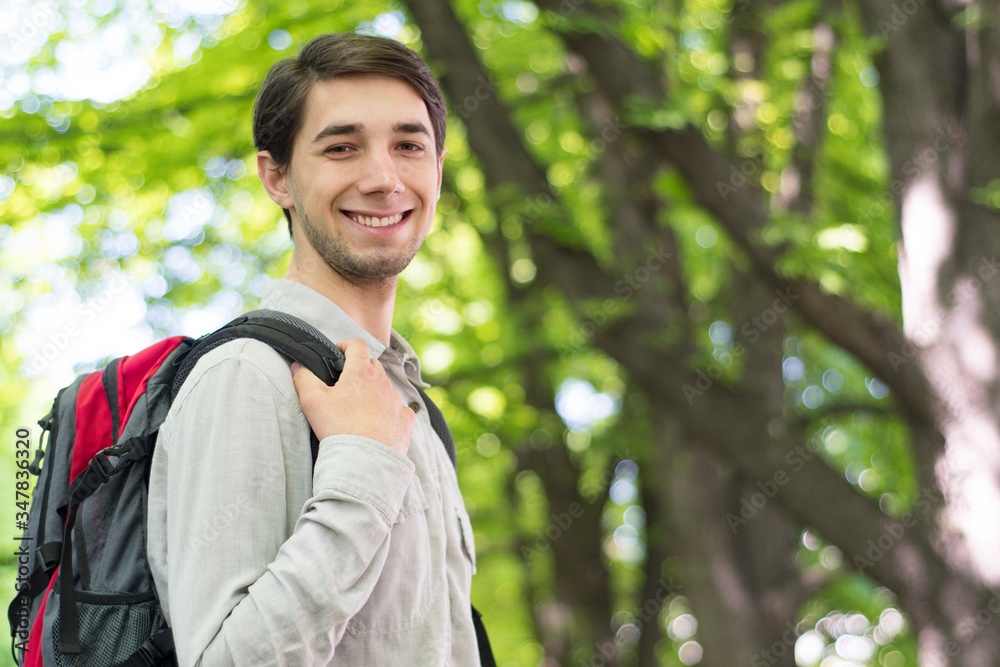 A beautiful young caucasian man with a backpack stands in the park and smiles. A beautiful young caucasian man with a backpack stands in the park and smiles.