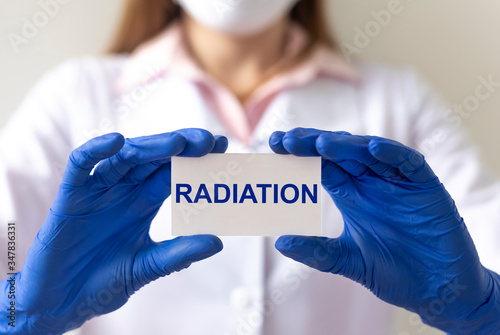 RADIATION text on white paper card in hands in blue protective gloves on white background with bluuring doctor photo